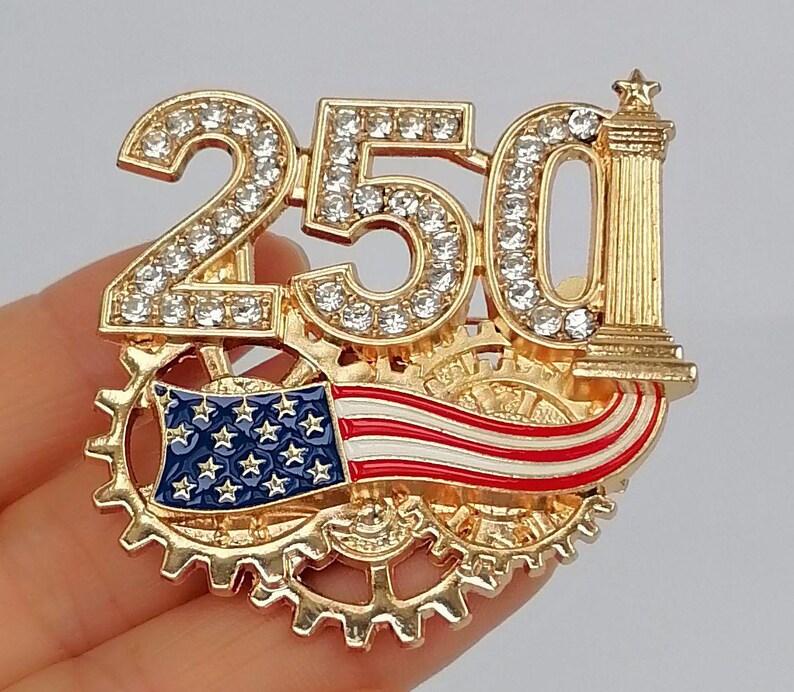 May include: Gold-toned brooch featuring the number "250" encrusted with clear rhinestones. The design includes a miniature American flag and a decorative column. The brooch is set against a backdrop of interlocking gears.