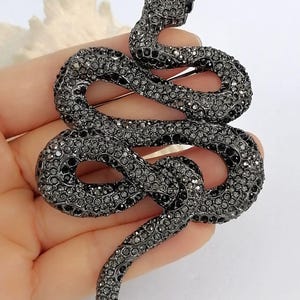 May include: A dark grey snake brooch, covered in small, sparkling crystals. The snake is coiled in a decorative pattern, with its head raised. This brooch is a fashion accessory, perfect for adding a touch of glamour to any outfit.