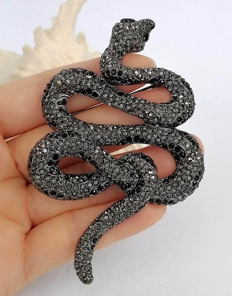 May include: A dark grey snake brooch, covered in small, sparkling crystals. The snake is coiled in a decorative pattern, with its head raised. This brooch is a fashion accessory, perfect for adding a touch of glamour to any outfit.