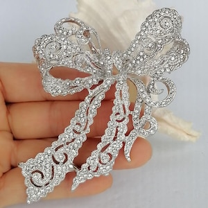 May include: A silver-toned bow brooch, encrusted with numerous clear crystals. The brooch features an openwork design with intricate details, creating a sparkling and elegant accessory. The bow's tails extend downwards, adding to its graceful appearance.