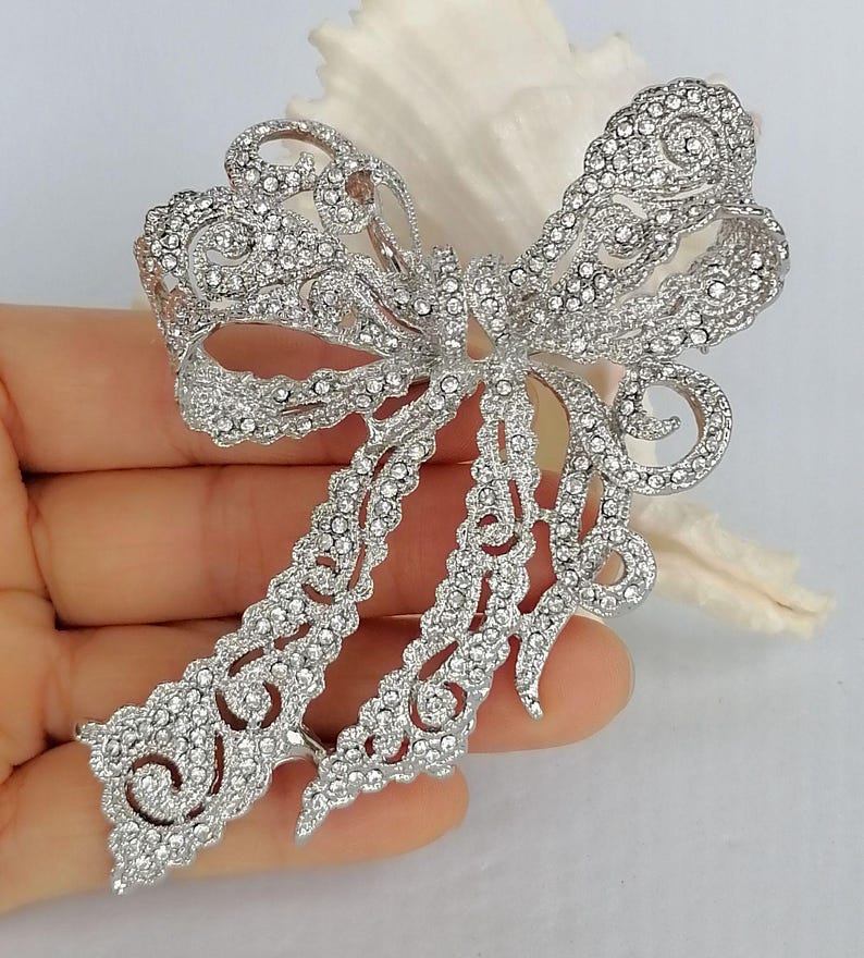 May include: A silver-toned bow brooch, encrusted with numerous clear crystals. The brooch features an openwork design with intricate details, creating a sparkling and elegant accessory. The bow's tails extend downwards, adding to its graceful appearance.