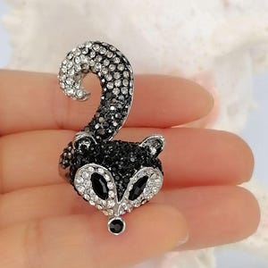 K&ouml;nnte beinhalten: Ein Ring in Form eines Waschb&auml;renkopfes mit geschwungenem Schwanz, besetzt mit schwarzen und klaren Kristallen. Das Waschb&auml;rengesicht hat Augen aus schwarzen Kristallen und eine kleine schwarze Kristallnase, w&auml;hrend Schwanz und Gesichtskontur gl&auml;nzen.