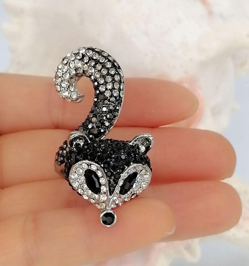 K&ouml;nnte beinhalten: Ein Ring in Form eines Waschb&auml;renkopfes mit geschwungenem Schwanz, besetzt mit schwarzen und klaren Kristallen. Das Waschb&auml;rengesicht hat Augen aus schwarzen Kristallen und eine kleine schwarze Kristallnase, w&auml;hrend Schwanz und Gesichtskontur gl&auml;nzen.