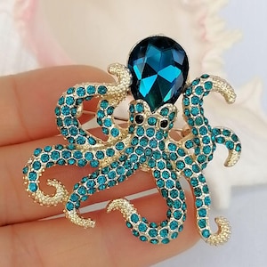 K&ouml;nnte beinhalten: Goldfarbene Oktopus-Brosche mit t&uuml;rkisfarbenen Strasssteinen. Der Oktopus hat einen gro&szlig;en, facettierten, t&uuml;rkisfarbenen Edelsteinkopf und schwarze Augen. Ein dekoratives Accessoire.