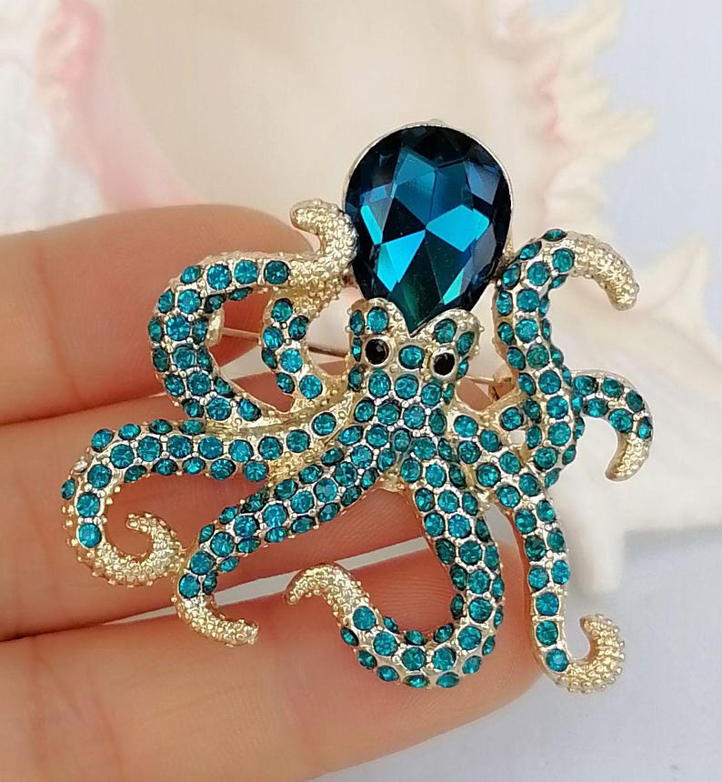 K&ouml;nnte beinhalten: Goldfarbene Oktopus-Brosche mit t&uuml;rkisfarbenen Strasssteinen. Der Oktopus hat einen gro&szlig;en, facettierten, t&uuml;rkisfarbenen Edelsteinkopf und schwarze Augen. Ein dekoratives Accessoire.