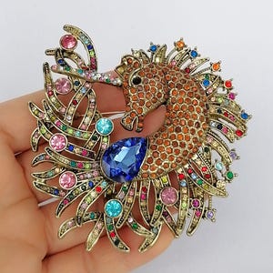 May include: A seahorse brooch with a bronze-tone metal base, adorned with orange, brown, and multicolored rhinestones. The seahorse features a large blue teardrop-shaped gem and is embellished with pink, blue, and green accents.