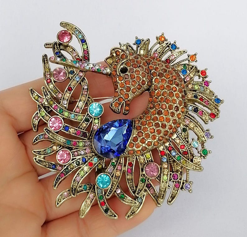 May include: A seahorse brooch with a bronze-tone metal base, adorned with orange, brown, and multicolored rhinestones. The seahorse features a large blue teardrop-shaped gem and is embellished with pink, blue, and green accents.