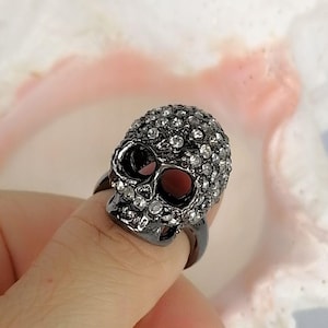 Op de afbeelding: Een ring in een donkere gunmetal-tint met een driedimensionaal schedelontwerp. Het oppervlak van de schedel is ingelegd met talloze kleine, heldere, reflecterende strass-steentjes die het voorhoofd, de jukbeenderen en de kaak bedekken.