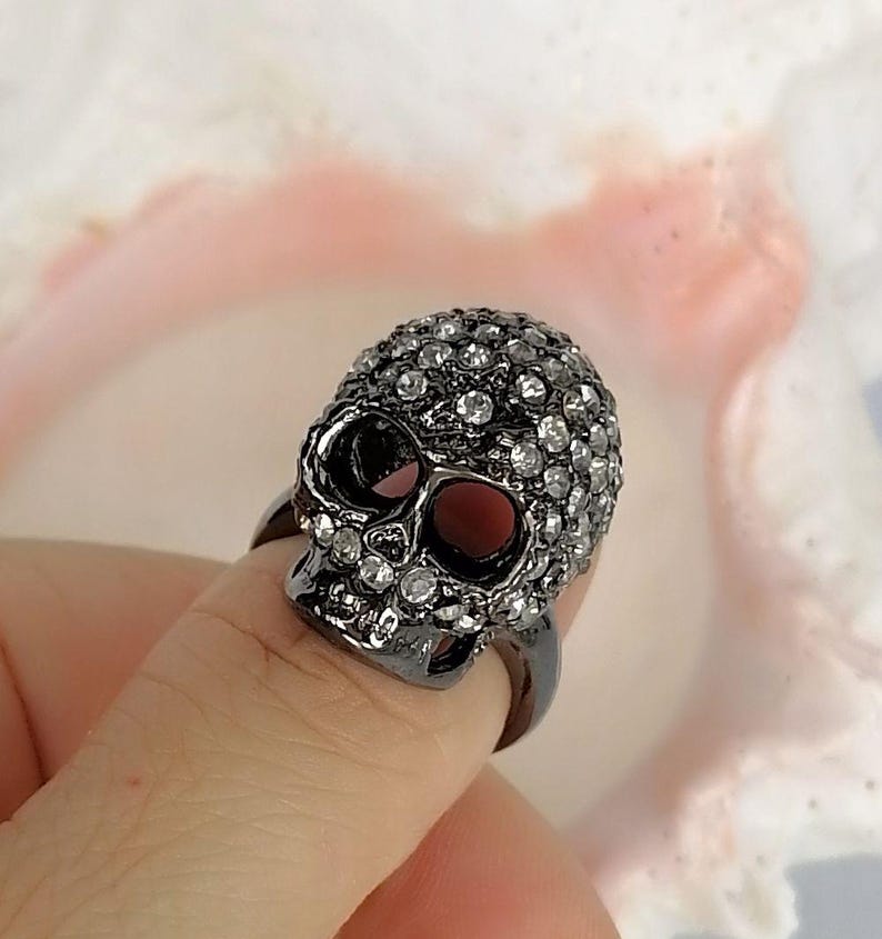 Op de afbeelding: Een ring in een donkere gunmetal-tint met een driedimensionaal schedelontwerp. Het oppervlak van de schedel is ingelegd met talloze kleine, heldere, reflecterende strass-steentjes die het voorhoofd, de jukbeenderen en de kaak bedekken.