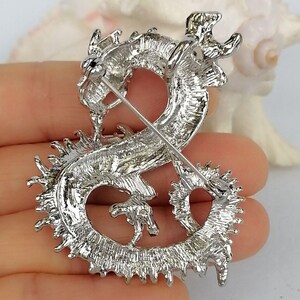 Puede incluir: Broche de drag&oacute;n plateado con detalles intrincados. El drag&oacute;n est&aacute; en una pose curvada, con escamas texturizadas y un cierre de alfiler en la parte posterior. El broche tiene un acabado met&aacute;lico brillante.