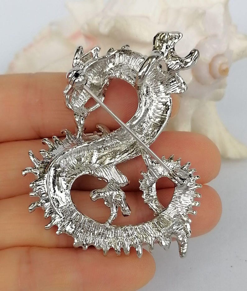 Puede incluir: Broche de drag&oacute;n plateado con detalles intrincados. El drag&oacute;n est&aacute; en una pose curvada, con escamas texturizadas y un cierre de alfiler en la parte posterior. El broche tiene un acabado met&aacute;lico brillante.