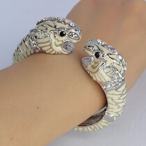 Può includere: Un bracciale rigido color argento con due teste di tigre contrapposte. Le teste sono rifinite con smalto color crema, dettagli neri per gli occhi e strass trasparenti lungo la parte superiore delle teste e sui collari delle figure.