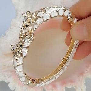 May include: A gold-tone hinged bangle bracelet featuring two giraffe heads meeting at the center. The bracelet is decorated with white enamel patches in a giraffe-like pattern and small clear stones along the top edge of the band.