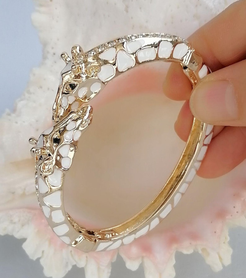May include: A gold-tone hinged bangle bracelet featuring two giraffe heads meeting at the center. The bracelet is decorated with white enamel patches in a giraffe-like pattern and small clear stones along the top edge of the band.