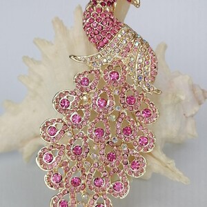 May include: A gold-toned peacock brooch with pink and clear crystal accents. The brooch features a detailed design with a fanned tail and a raised head. The crystals are set in a gold-colored metal, creating a sparkling effect.