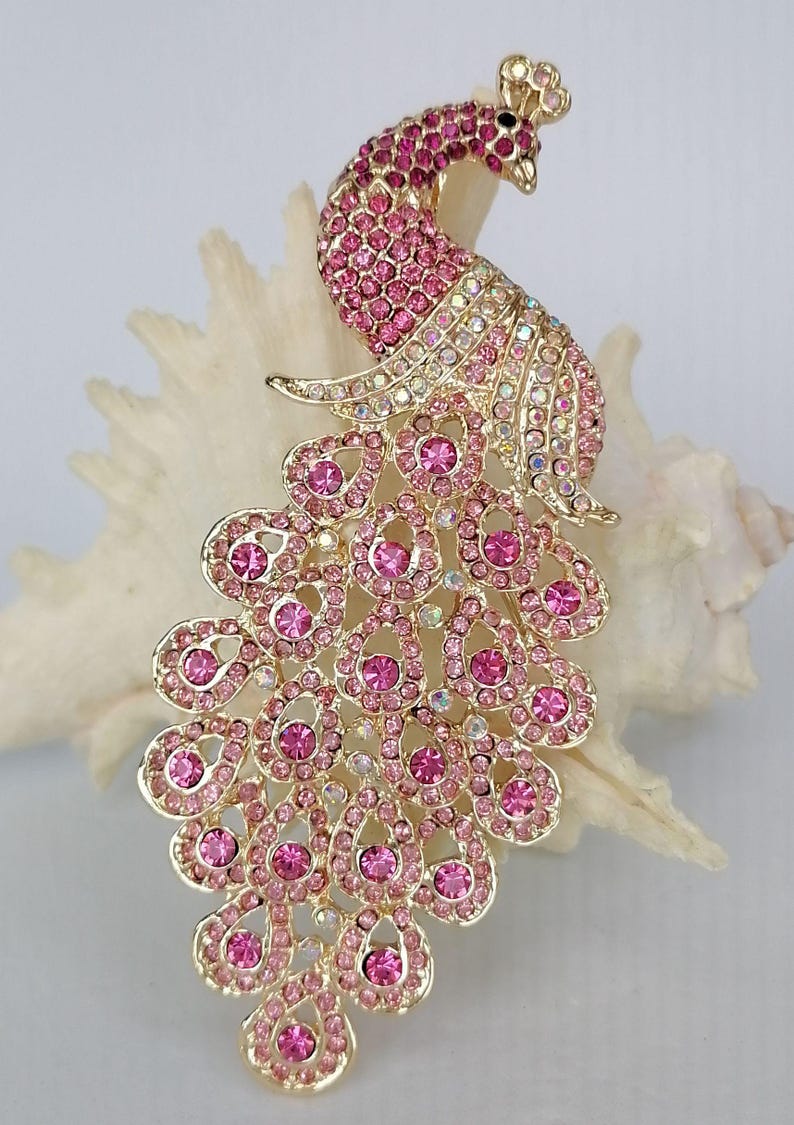 May include: A gold-toned peacock brooch with pink and clear crystal accents. The brooch features a detailed design with a fanned tail and a raised head. The crystals are set in a gold-colored metal, creating a sparkling effect.