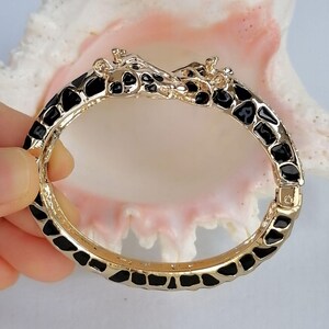 May include: A gold-tone hinged bangle bracelet featuring a giraffe motif at the clasp. The bracelet is decorated with a black enamel pattern that mimics giraffe spots, covering the entire outer surface of the metal band in a continuous design.