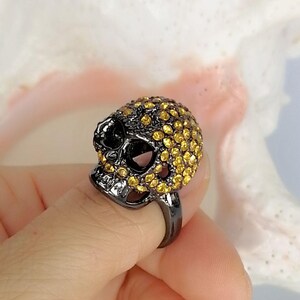 Op de afbeelding: Een ring met een schedelvormig middendeel van donkergrijs metaal. Het oppervlak van de schedel is dicht bezet met talloze kleine, ronde, goudgele kristallen die de schedel en de gezichtsstructuur van het object bedekken.