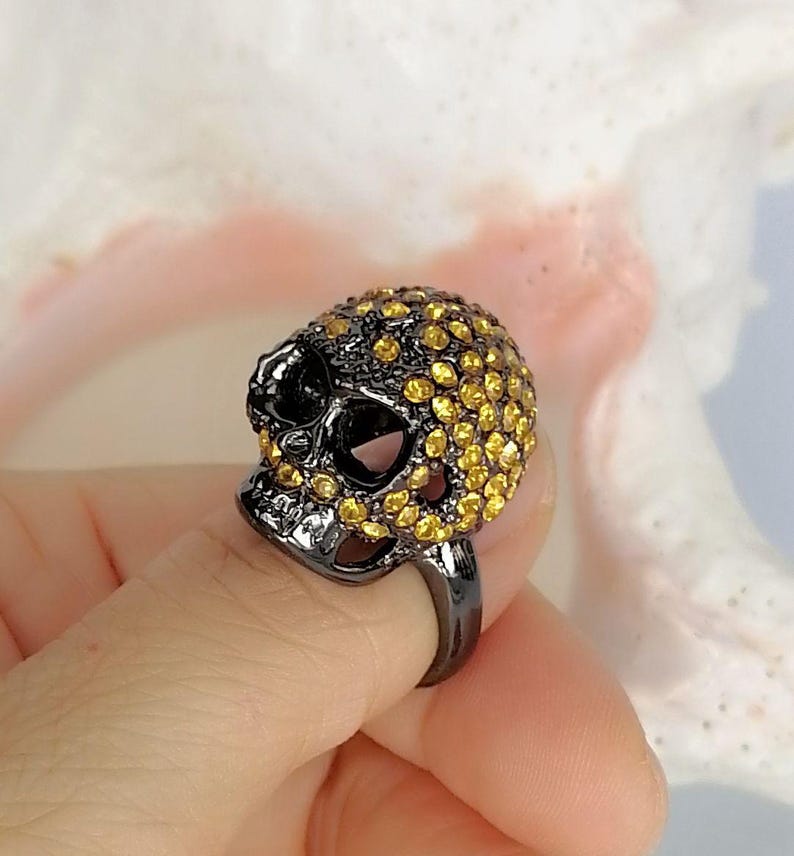 Op de afbeelding: Een ring met een schedelvormig middendeel van donkergrijs metaal. Het oppervlak van de schedel is dicht bezet met talloze kleine, ronde, goudgele kristallen die de schedel en de gezichtsstructuur van het object bedekken.