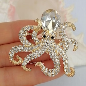 K&ouml;nnte beinhalten: Goldfarbene Oktopus-Brosche, besetzt mit klaren Kristallen. Der Oktopus hat einen gro&szlig;en, facettierten Kristallkopf und schwarze Augen. Ein detailliertes, funkelndes Accessoire.