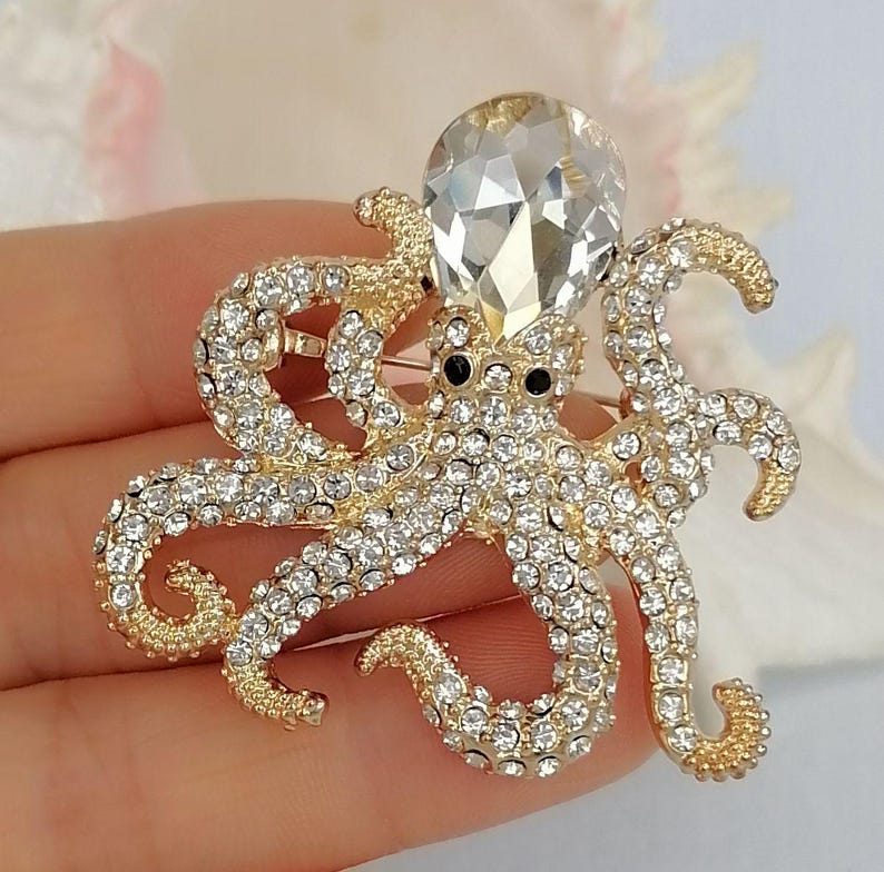 K&ouml;nnte beinhalten: Goldfarbene Oktopus-Brosche, besetzt mit klaren Kristallen. Der Oktopus hat einen gro&szlig;en, facettierten Kristallkopf und schwarze Augen. Ein detailliertes, funkelndes Accessoire.