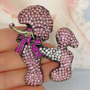 May include: A poodle-shaped brooch covered in light pink and clear rhinestones. The dog figure features a dark metal finish, a bright pink rhinestone bow around its neck, and a single dark pink rhinestone eye, creating a detailed animal design.