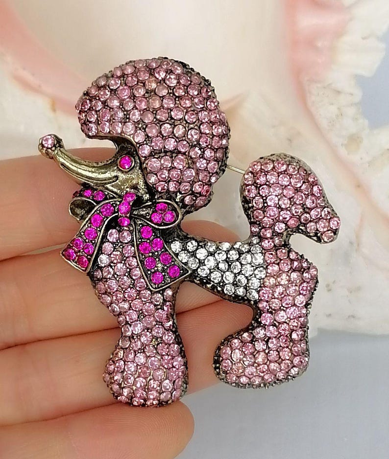 May include: A poodle-shaped brooch covered in light pink and clear rhinestones. The dog figure features a dark metal finish, a bright pink rhinestone bow around its neck, and a single dark pink rhinestone eye, creating a detailed animal design.