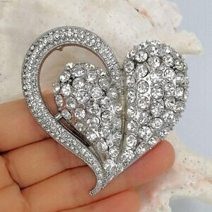 May include: Silver-toned heart-shaped brooch encrusted with clear rhinestones. The brooch features a large heart shape with a smaller heart cutout, and is embellished with numerous round, sparkling crystals. The brooch is held in a hand.