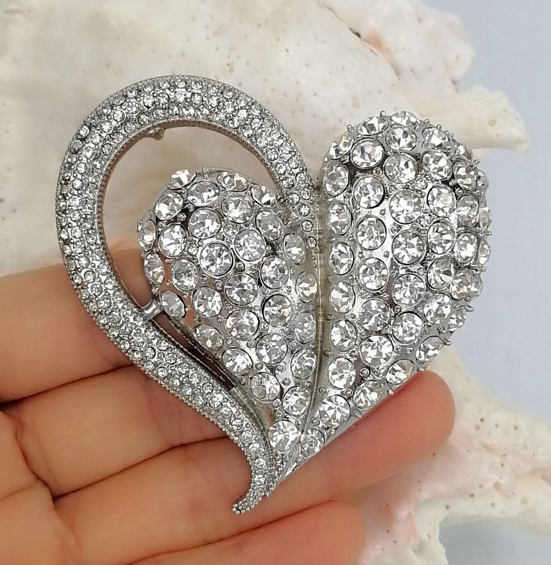 May include: Silver-toned heart-shaped brooch encrusted with clear rhinestones. The brooch features a large heart shape with a smaller heart cutout, and is embellished with numerous round, sparkling crystals. The brooch is held in a hand.
