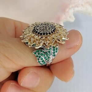 K&ouml;nnte beinhalten: Ein sonnenblumenf&ouml;rmiger Ring mit einem dunklen Zentrum aus schwarzen Steinen, goldfarbenen Bl&uuml;tenbl&auml;ttern und einem silberfarbenen Blattakzent, der mit kleinen, leuchtend gr&uuml;nen Steinen besetzt ist. Der Ring hat ein detailliertes Design.