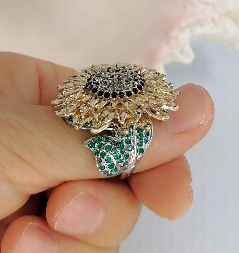 K&ouml;nnte beinhalten: Ein sonnenblumenf&ouml;rmiger Ring mit einem dunklen Zentrum aus schwarzen Steinen, goldfarbenen Bl&uuml;tenbl&auml;ttern und einem silberfarbenen Blattakzent, der mit kleinen, leuchtend gr&uuml;nen Steinen besetzt ist. Der Ring hat ein detailliertes Design.