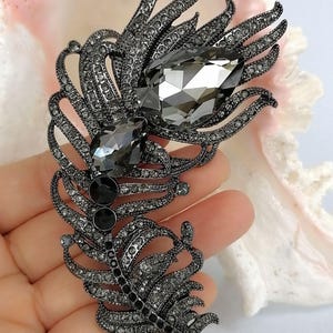 May include: A feather-shaped brooch crafted from dark gunmetal-toned metal, densely encrusted with small gray crystals. The design features two prominent, large, faceted teardrop-shaped gray stones positioned along the central spine of the feather.