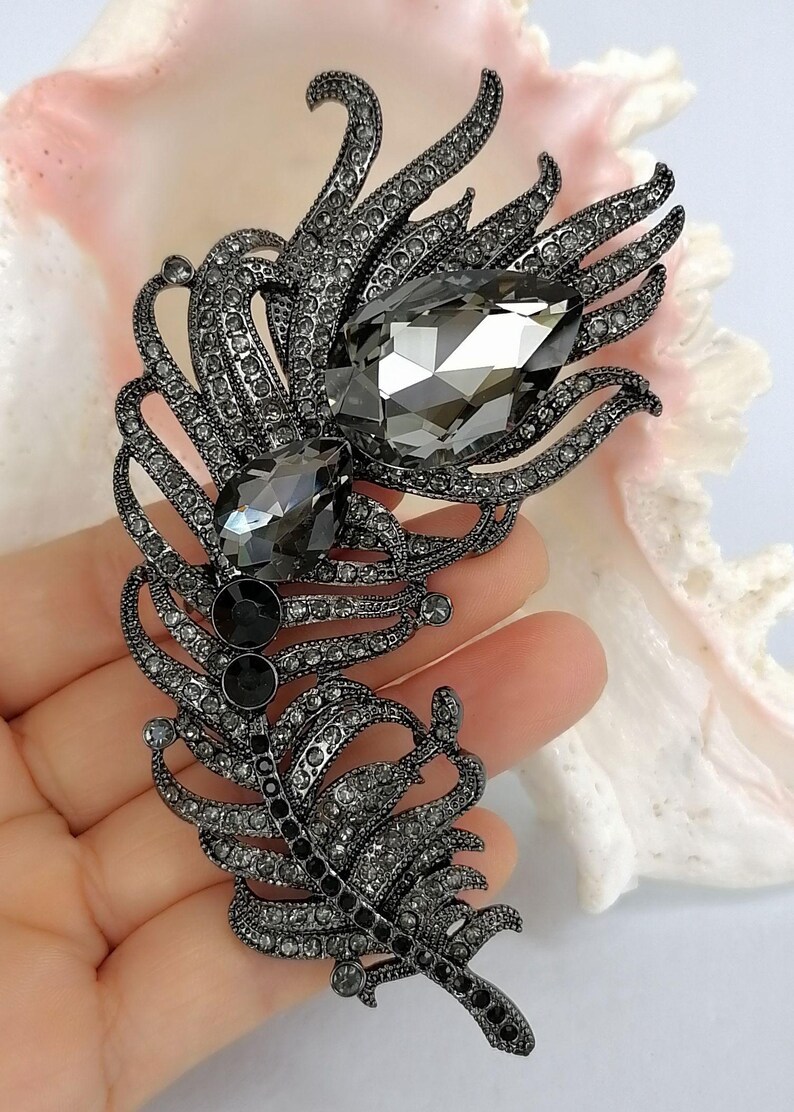 May include: A feather-shaped brooch crafted from dark gunmetal-toned metal, densely encrusted with small gray crystals. The design features two prominent, large, faceted teardrop-shaped gray stones positioned along the central spine of the feather.