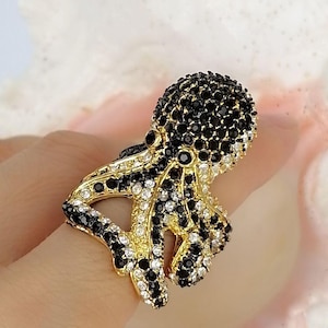 Peut inclure: Une bague dor&eacute;e en forme de pieuvre, dont la t&ecirc;te et les tentacules sont incrust&eacute;s de nombreuses petites pierres &eacute;tincelantes noires et blanches transparentes. La base m&eacute;tallique est visible entre les pierres, formant la structure de l'animal.