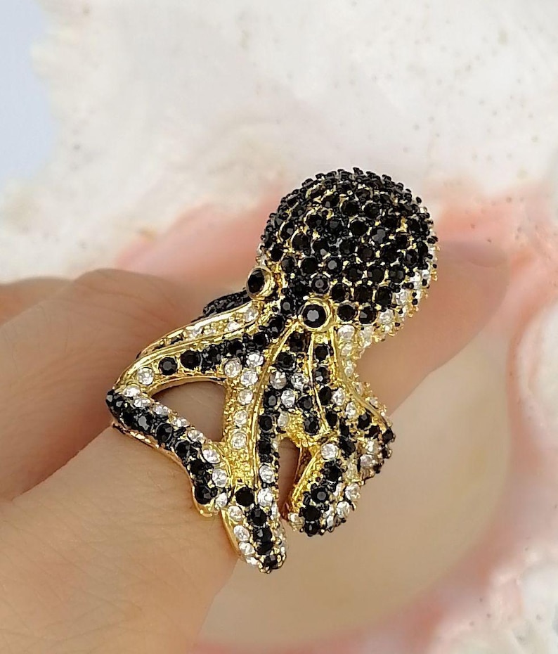 Peut inclure: Une bague dor&eacute;e en forme de pieuvre, dont la t&ecirc;te et les tentacules sont incrust&eacute;s de nombreuses petites pierres &eacute;tincelantes noires et blanches transparentes. La base m&eacute;tallique est visible entre les pierres, formant la structure de l'animal.