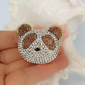 May include: A panda-shaped brooch covered in sparkling clear and amber-colored rhinestones. The panda face features two amber-colored patches around the eyes, amber-colored ears, and a small black bead representing the nose, all set in gold metal.