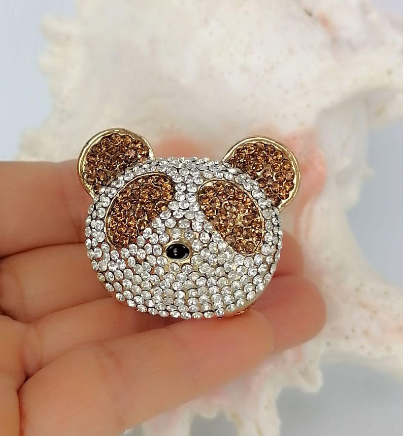 May include: A panda-shaped brooch covered in sparkling clear and amber-colored rhinestones. The panda face features two amber-colored patches around the eyes, amber-colored ears, and a small black bead representing the nose, all set in gold metal.