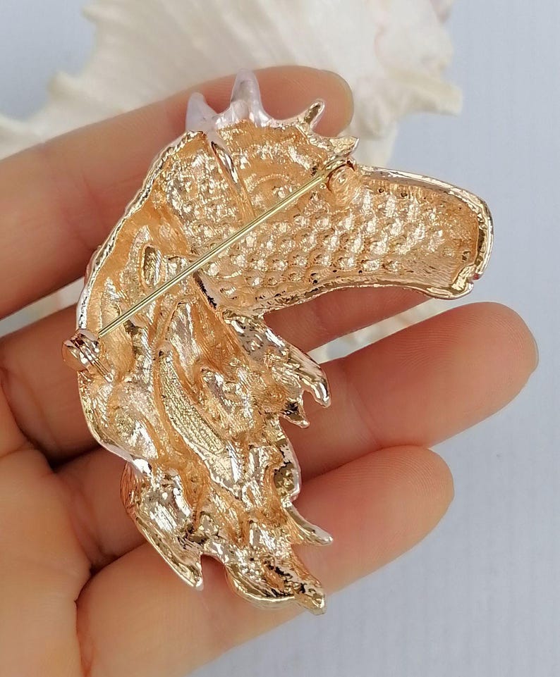 K&ouml;nnte beinhalten: Eine goldfarbene Pferdekopfbrosche. Die Brosche ist mit strukturierten Oberfl&auml;chen und einem Nadelverschluss auf der R&uuml;ckseite versehen. Der Pferdekopf hat eine M&auml;hne und ein detailliertes Gesicht. Die Brosche ist wahrscheinlich aus Metall.