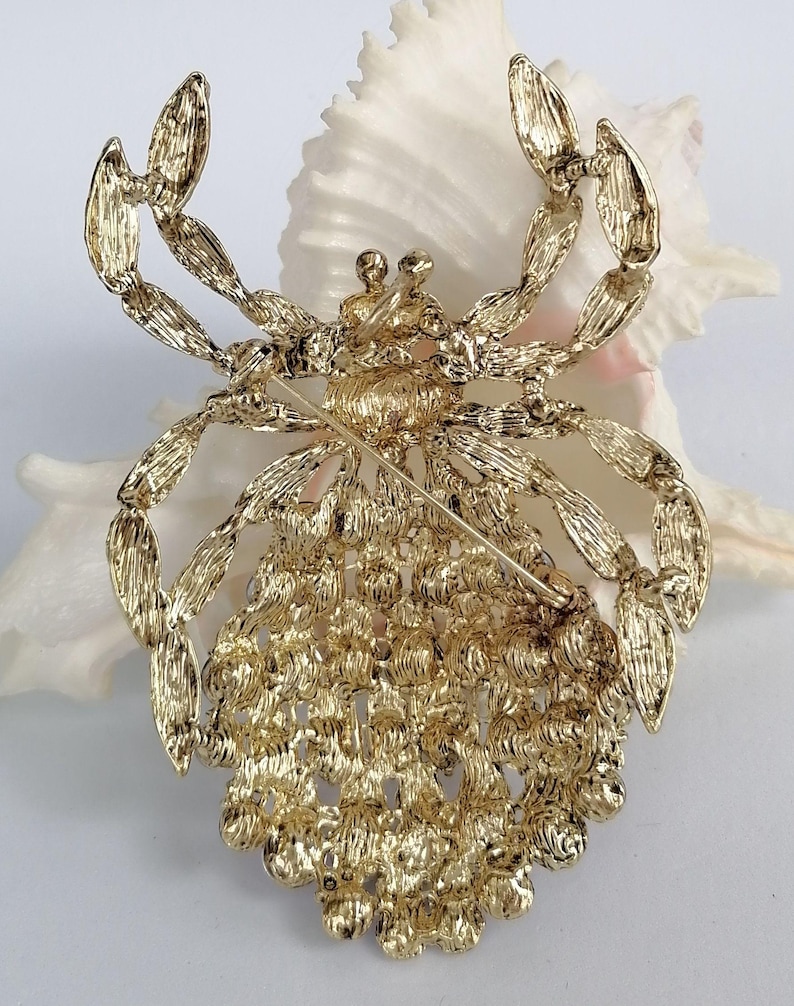May include: A gold-coloured metal crab brooch with detailed craftsmanship. The crab's body features small, circular decorations, while its legs and claws are finely shaped. The brooch has a pin fastening on the reverse, designed for attaching to garments or accessories.