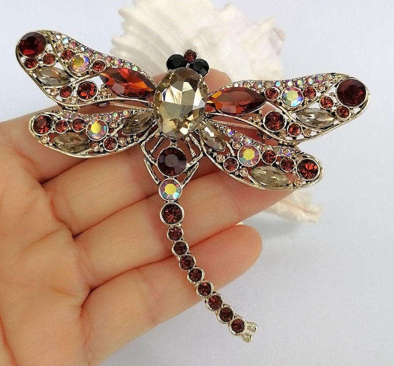 May include: A dragonfly brooch with a gold-tone metal frame, decorated with red, brown, and clear crystals. The dragonfly's body has a large, faceted amber crystal. The wings are embellished with smaller crystals.