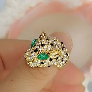 May include: A gold-toned ring shaped like a leopard head, featuring clear white rhinestones and black stones covering the surface, with two marquise-cut green stones serving as the eyes of the feline figure in a detailed metal setting.