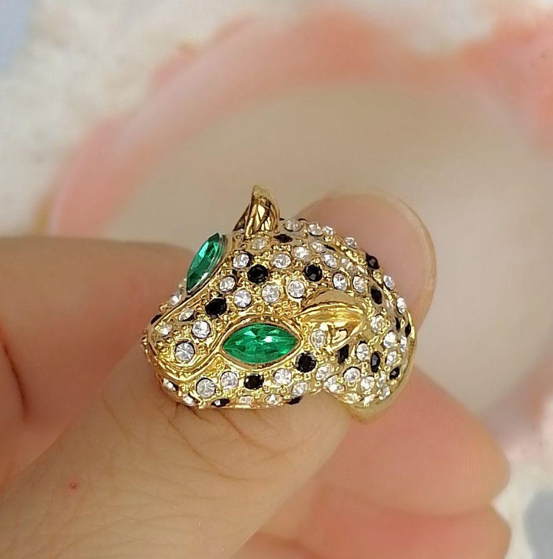 May include: A gold-toned ring shaped like a leopard head, featuring clear white rhinestones and black stones covering the surface, with two marquise-cut green stones serving as the eyes of the feline figure in a detailed metal setting.