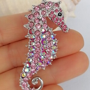 May include: A silver-toned seahorse brooch encrusted with pink and iridescent crystals. The seahorse has a detailed design with a curled tail and a black eye. The brooch is a decorative accessory.