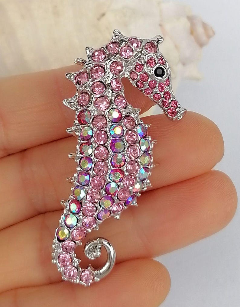 May include: A silver-toned seahorse brooch encrusted with pink and iridescent crystals. The seahorse has a detailed design with a curled tail and a black eye. The brooch is a decorative accessory.
