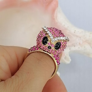 May include: A gold-toned ring featuring a three-dimensional owl design covered in small pink crystals. The owl has two large, smooth black oval eyes and a row of clear white crystals forming a brow line across the top of its head.