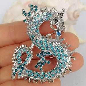 Puede incluir: Broche de drag&oacute;n plateado con pedrer&iacute;a turquesa. El drag&oacute;n presenta un cuerpo texturizado y un dise&ntilde;o intrincado. Un accesorio decorativo para ropa o bolsos.