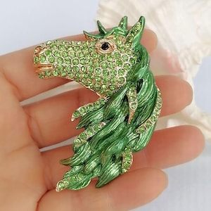 K&ouml;nnte beinhalten: Eine gr&uuml;ne und goldene Pferdekopfbrosche. Der Pferdekopf ist mit gr&uuml;nen Strasssteinen besetzt und hat eine flie&szlig;ende gr&uuml;ne M&auml;hne. Die Brosche wird in einer Hand gehalten.