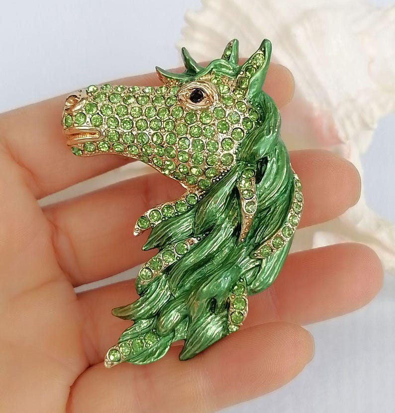 K&ouml;nnte beinhalten: Eine gr&uuml;ne und goldene Pferdekopfbrosche. Der Pferdekopf ist mit gr&uuml;nen Strasssteinen besetzt und hat eine flie&szlig;ende gr&uuml;ne M&auml;hne. Die Brosche wird in einer Hand gehalten.