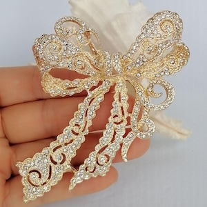 May include: A gold-toned bow brooch, encrusted with clear crystals. The brooch has an openwork design with intricate details, including swirling patterns and delicate edges. The bow's shape is clearly defined, with two loops and two flowing tails.
