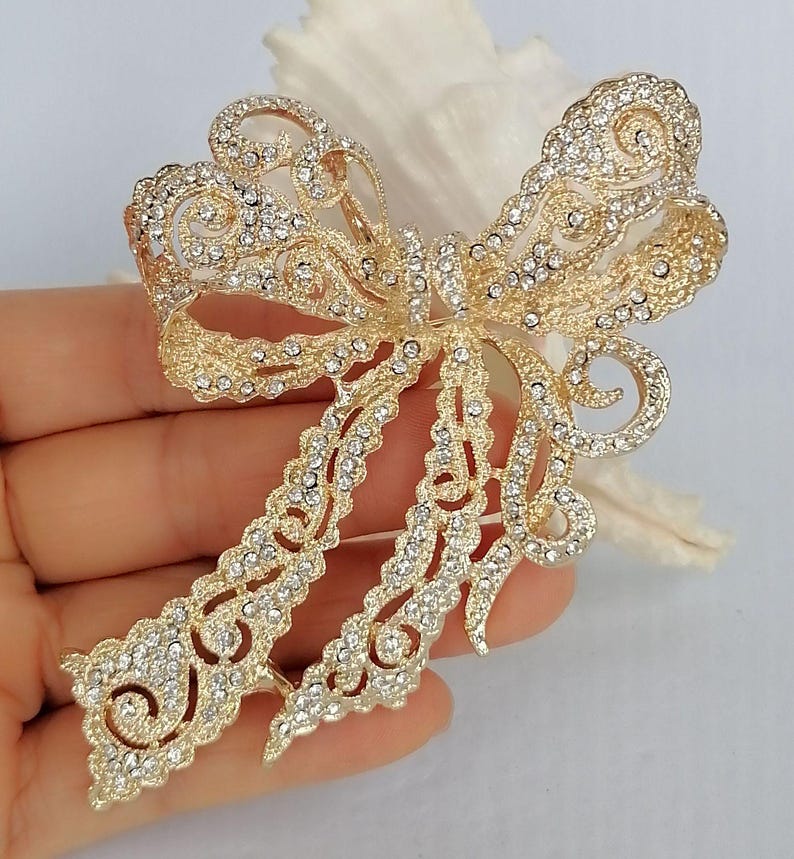 May include: A gold-toned bow brooch, encrusted with clear crystals. The brooch has an openwork design with intricate details, including swirling patterns and delicate edges. The bow's shape is clearly defined, with two loops and two flowing tails.
